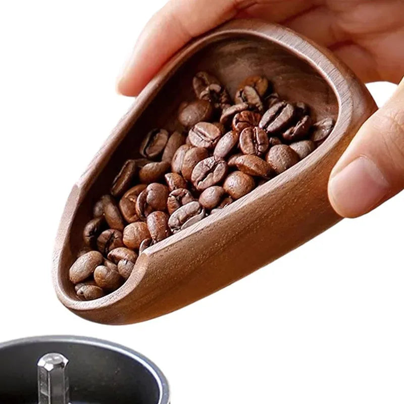 Wooden scoop filled with coffee beans held by a hand, with a blurred coffee grinder in the background.